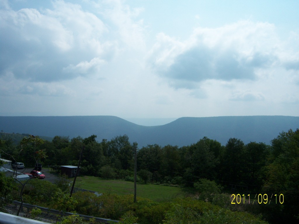 The forground is green grass and a stand of darker green trees. Beyond the trees a good many miles away is a mountain range. There is a very conspicuous dip in the otherwise straight line of a mountain ridge in the shape of a saddle; hence the name "Saddle Mountain."