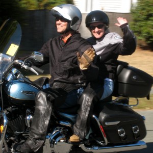Ed & Marilee riding on a Harley-Davidson Road King motorcycle. As they pass the camera, they smile and wave but Ed keeps his eyes on the road.