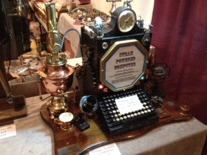 A steam powered computer. An interesting example of employing improbable anachronisms. The odd looking but recognizable computing device sports a teletypewriter keyboard, a telegraph key, an anniversary clock, and a teapot (for the steam power of course).