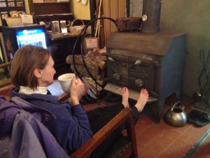 In photo. A skier sits in a rocking chair in front of the wood burning stove. Her bare feet with painted toenails are warming near the stove as she sips hot cocoa. End photo.