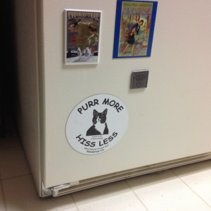 In photo. A large round magnetic sticker is seen on the lower left hand corner of the refrigerator door. The phrase around the circle of the sticker are "Purr more. Hiss less." There is a calm cat in the center of the words with whose markings are quite similar to Holly and Wednesday. End photo.