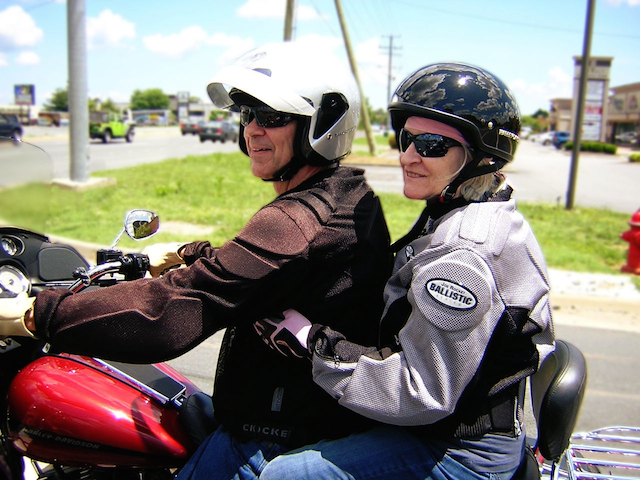 In photo. Ed and Marilee are on their motorcycle waiting at a stoplight. Both are looking to the left and smiling for the camera. End photo.