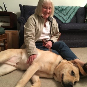 Marilee is on the floor leaning back on a couch. Ryder, out of harness, is laying next to her on the right.