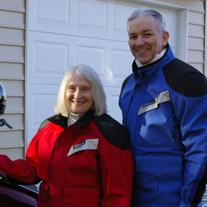 Image of Marilee and Ed suited up in winter motorcycle riding attire. Not leather but exotic 21st century materials designed to protect in the event of a spill. But also quite stylish. Marilee is in red while Ed is in blue.