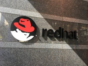 In photo. The logo of Red Hat is a red fedora worn by a featureless neuter gender head. Next to the graphic are the lower case letters which spell the single word "redhat." Out of photo.