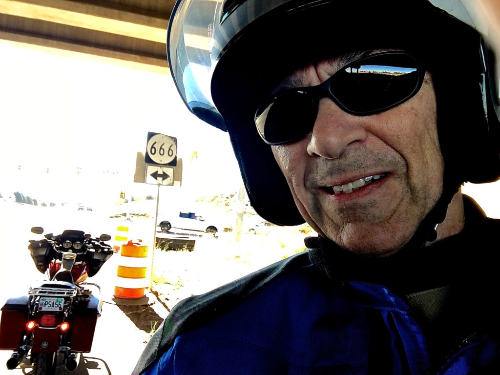 In photo. A selfie of Ed the rider. Over his right shoulder the viewer is able to see the motorcycle parked near a road sign indicating "Route 666." End photo.