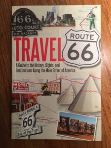 In photo. Image of book cover. Title is "Travel Route 66." Images on the cover include postcard photos of attractions along and maps of the route. End photo.
