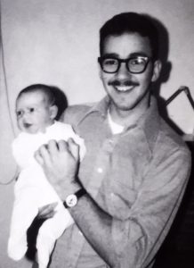 In Photo: A much younger Ed is holding an infant Tabitha. She in feety pajamas and looking away from the camera while Ed holds her upright and smiles for the camera. Out of Photo.