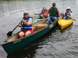 In Photo: Marilee, Ryder, and Ed are in a canoe on the water. Along side on left and right are two other Sports For Health participants in kayaks. Ed is holding the kayaks to keep them close for the photo op of the tiny flotilla. Out of Photo.