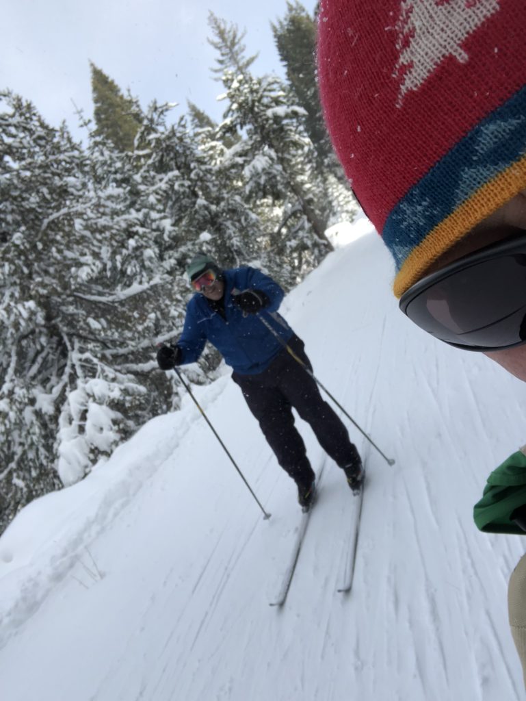 In photo. Larry is seen over the shoulder of Ed who has used the selfie feature of his smart phone while skiing down the hill to the valley. Larry is double poling and is captured mid stride as he begins another downward push. Out of photo.