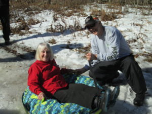 In photo. They are posed for the camera. Marilee sits in the tire tube snow toy while Ed prepares to propel her down Marty's Bump. Out of photo.