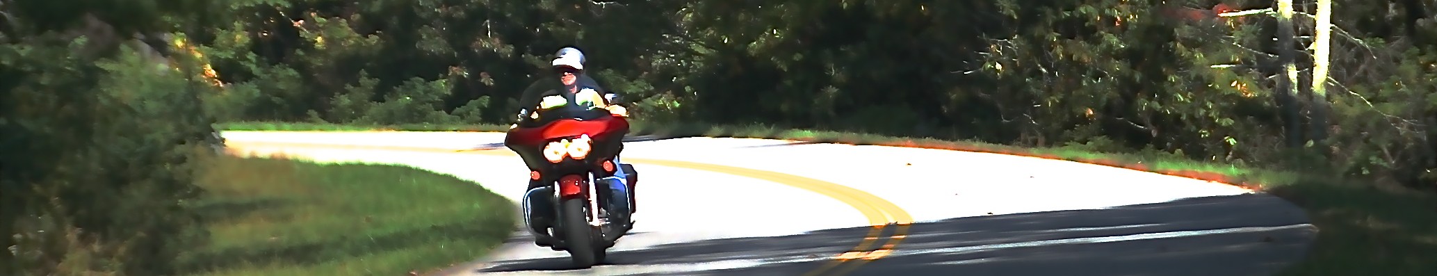 In photo. A red Harley-Davidson Road Glide motorcycle is seen rounding a turn on a two-lane tree lined country road. End photo.