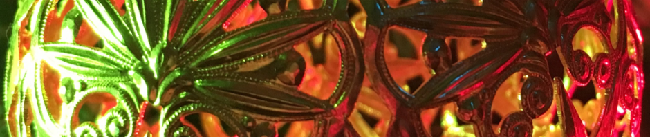 The banner photo says "It's Christmas" in the form of a brass tree ornament. Half of the lattice work sphere is lit by a green light and the other half a red light. The openings in the design of the ball allow the viewer to see through to the other side. The design is composed of a series of eight point stars and intricate lace work connecting a shell pattern with vines and structural swirls of delicate metal.