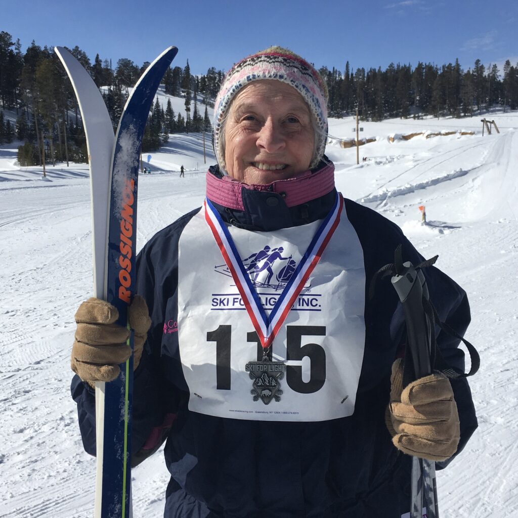 Marilee holds her skis and poles while wearing her event medal. In the background is seen the snowy valley and ski tracks of the main trail.