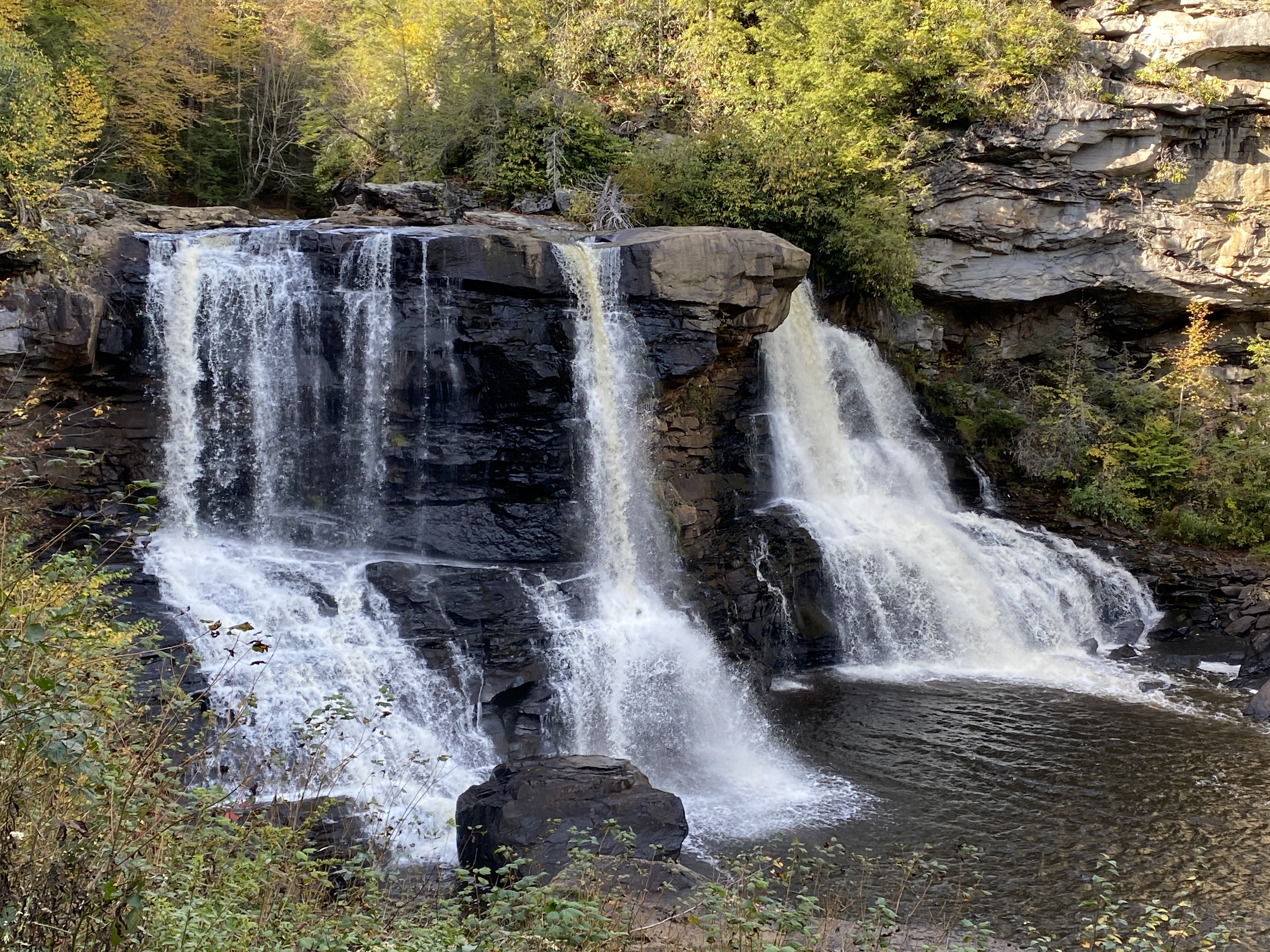 IN PHOTO. This is quintessential postcard photo of the falls with a goodly flow of water breaking over the equivalent of a four story building. The water and rocks are framed by trees of various colors dappled in sunlight. OUT OF PHOTO.